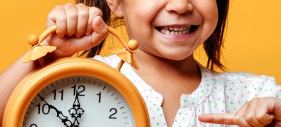 Smiling little girl child holding clock alarm. Photo of smiling little girl child standing isolated over yellow background. Looking camera holding clock alarm.