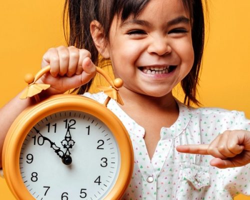 Smiling little girl child holding clock alarm. Photo of smiling little girl child standing isolated over yellow background. Looking camera holding clock alarm.